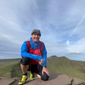 Alan Stone smiling in the sun with Pen y Fan and Corn Du in the background